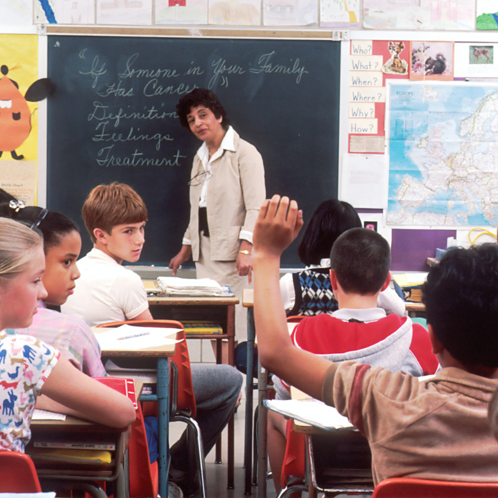 Student raises a hand to ask a question in a classroom while the teacher stands at the board.