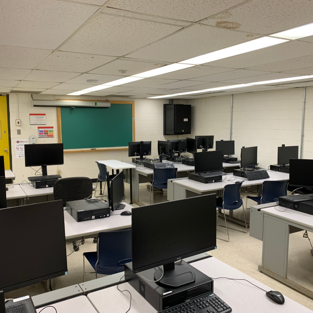 A computer lab with multiple workstations. Each desk has a desktop computer and monitor, arranged in rows facing a central aisle. At the front, there's a teacher's desk with a computer and a blank green chalkboard on the wall above. The room has fluorescent lighting and a white tiled ceiling. There's no projector visible. The perfect environment for the RODE Wireless GO II.
