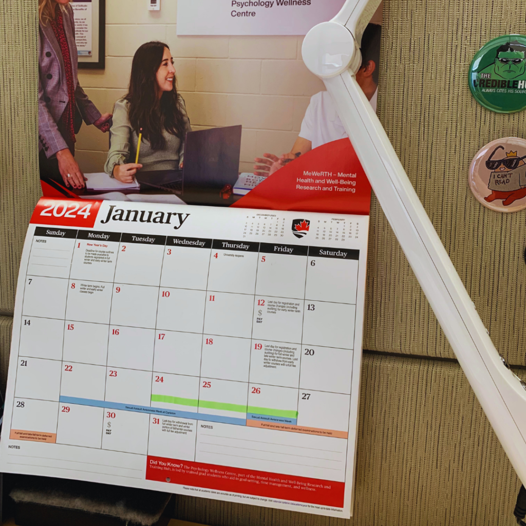 A corner of an office space with a wall-mounted calendar showing January 2024. The calendar is opened to a page with a vibrant red banner for January, below which is a stock image of a smiling woman at a desk talking to someone out of frame. Above the calendar is a poster labeled 'Psychology Wellness Centre.' To the right of the calendar, pinned to a fabric cubicle wall, are a couple of whimsical pins.