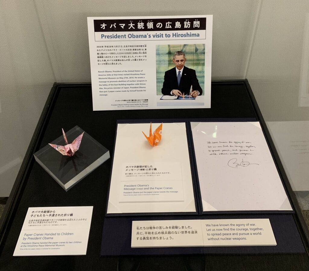 The image shows a display dedicated to President Obama's visit to Hiroshima. The central focus is on two origami paper cranes placed in a display case, one pink and one orange, with accompanying text indicating that President Obama handed these cranes to children during his visit to the Hiroshima Peace Memorial Museum. Above this, a larger text panel and photo show President Obama signing a message. The message is displayed below the photo and reads, "We have known the agony of war. Let us now find the courage, together, to spread peace and pursue a world without nuclear weapons." The text is in both English and Japanese, reflecting the bilingual nature of the exhibit and its international significance. The display is a poignant reminder of President Obama's historic visit to Hiroshima and his call for peace and disarmament. Like my own evolution, the U.S. too, evolves to a changing political landscape.