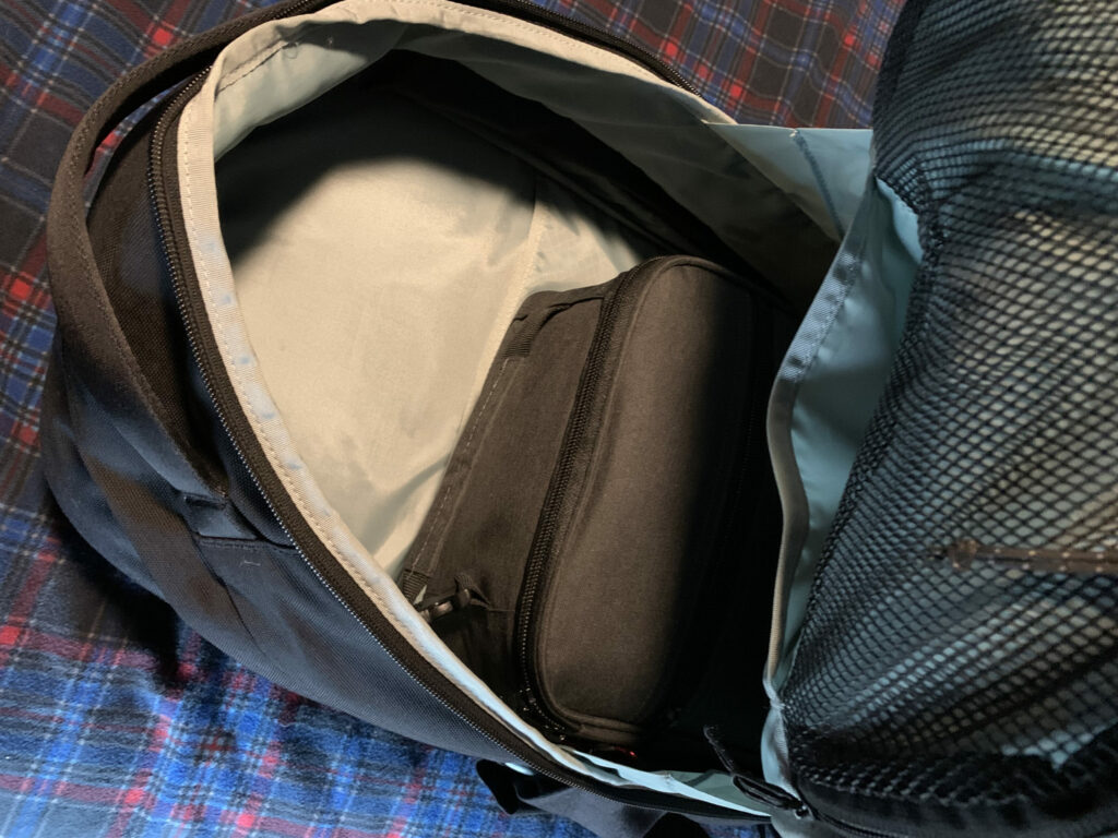 "An open black Timbuk2 backpack showing a packed gray Hap Tim medical supplies bag placed inside the main compartment. The interior of the backpack is light gray with a mesh pocket in the lid, all resting on a blue and red plaid fabric surface.