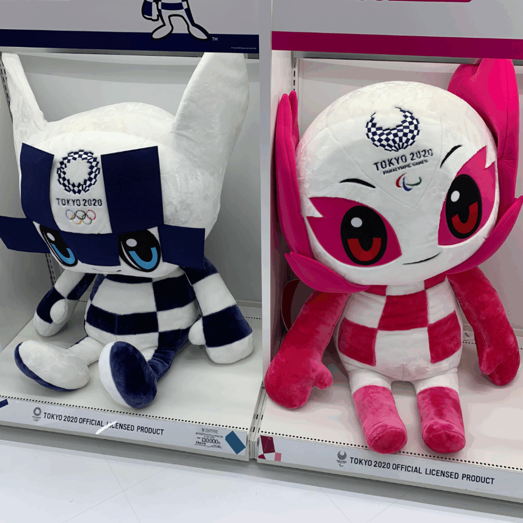 Two large plush toys representing the Tokyo 2020 Olympic and Paralympic Games mascots are displayed side by side on a white shelf. The left toy, with blue and white checkered patterns and large blue eyes, is the Olympic mascot Miraitowa. The right toy, with pink and white checkered patterns and vivid red eyes, is the Paralympic mascot Someity. Both are official licensed products.