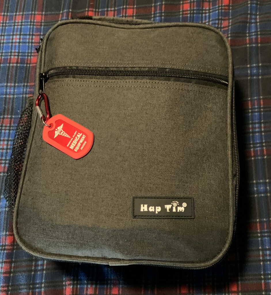 A closed gray Hap Tim travel bag resting on a blue and red plaid fabric surface. Attached to the bag’s zipper is a red medical alert tag indicating that medical equipment is inside.