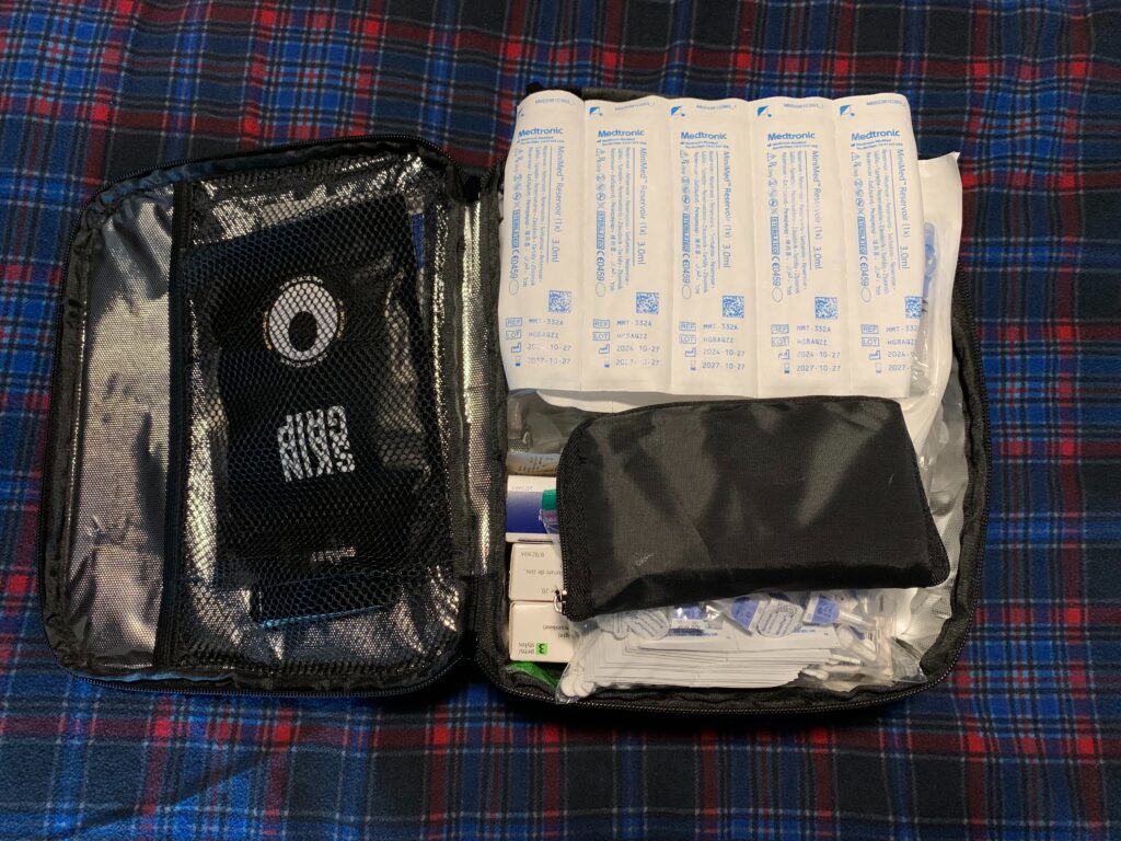 An open black travel case packed with medical supplies, including several Medtronic MiniMed reservoir packages neatly lined up, a black mesh pocket containing a circular medical device, and various small medical items organized underneath a small black pouch. The case is placed on a blue and red plaid fabric surface.