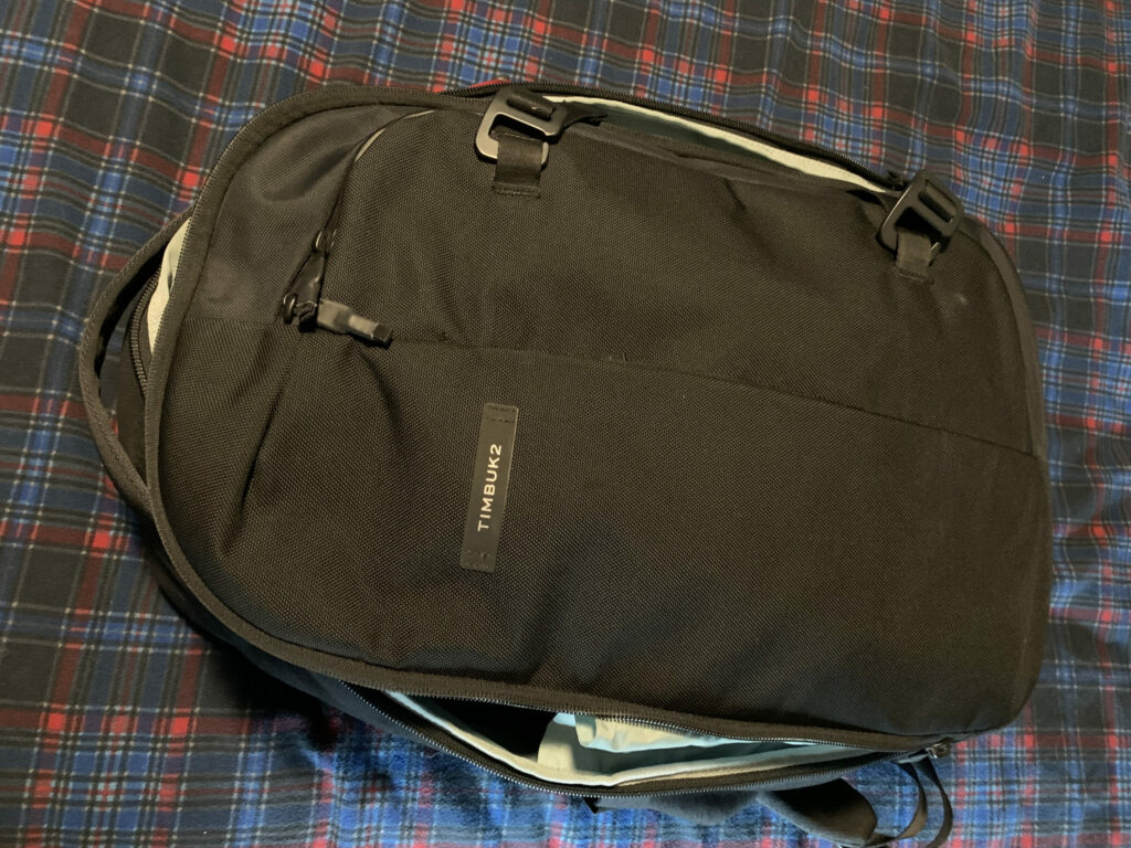 A closed black Timbuk2 backpack resting on a blue and red plaid fabric surface. The zippers are partially open, revealing a glimpse of the light gray interior.